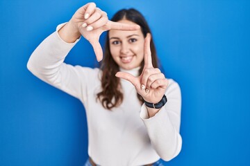 Sticker - Young hispanic woman standing over blue background smiling making frame with hands and fingers with happy face. creativity and photography concept.