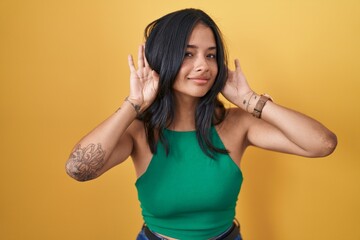 Canvas Print - Brunette woman standing over yellow background trying to hear both hands on ear gesture, curious for gossip. hearing problem, deaf