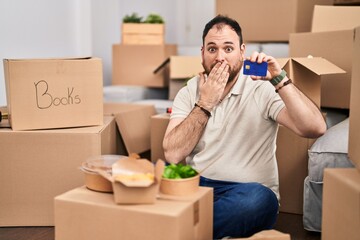 Wall Mural - Plus size hispanic man with beard moving to a new home holding credit card covering mouth with hand, shocked and afraid for mistake. surprised expression