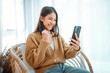 © oatawa - Excited young asian woman hold phone chatting in dating app feel happy sit on sofa at home, Closeup joyful female reading good news on phone. Surprised lady celebrating victory on phone