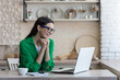 © Liubomir - A beautiful young woman communicates on a video call on a laptop, watches a video, a movie. Sitting at the kitchen table, looking at the screen, smiling.
