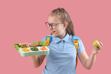 Happy little girl with backpack, lunchbox and apple on pink background