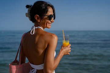 Wall Mural - a tanned girl holding a glass of orange juice and a bag in her hand stands on the sand against the backdrop of the sea