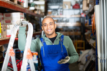 Positive warehouse worker stands next to stepladder and tool shelves in a hardware store