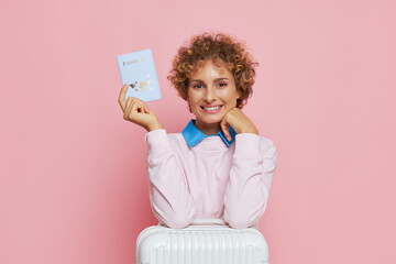 Wall Mural - Tourist girl wearing light pink sweatshirt showing passport with smile posing with white suitcase on pink background, happy journey concept, copy space