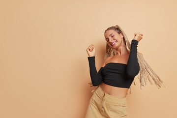 Wall Mural - Happy girl with long dreadlocks dressed in black long sleeve top dancing with hands up on light brown backdrop, happiness concept, copy space