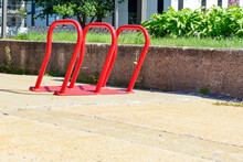 Multiple Large Red Metal Bicycling Hub Racks. The Curved Shape Of The Metal Stands Is Attached To The Edge Of A Brick Garden Wall. There Are Hosta Flowers And Grass In The Background.