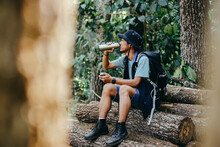 Tired Asian Hiker Man Sitting On Pile Of Log Wood, Drinking Water From A Bottle While Resting During Hiking In The Forest.
