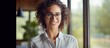 © HN Works - A confident young Caucasian woman stands at a window wearing a shirt and glasses holding a tablet smiling