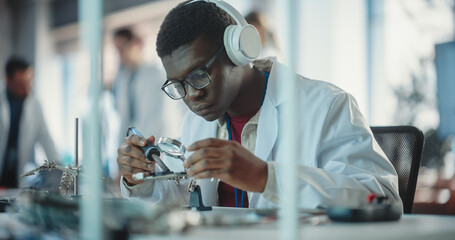 Poster - Portrait of Black Male Lab Specialist Soldering a Circuit Board, Using Headphones in an Industrial Company. Professional Man Listening to Music, Working on Technological Project For a Startup