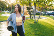 © Strelciuc - Confident young woman enjoying morning coffee in park holding yoga mat. Holding rolled mat and plastic cup of coffee. Having break after yoga class