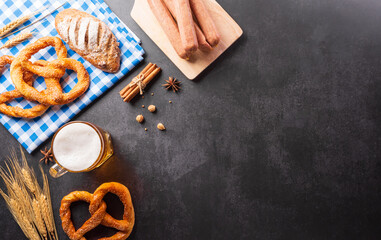 Wall Mural - Oktoberfest festival decoration symbols made from Pretzel loaf, beer, sausage, potato chips and Bavarian white and blue fabric on dark stone background.