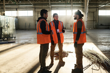 Leinwandbilder - Group of three young intercultural builders in workwear discussing working points at meeting while standing in warehouse or workshop