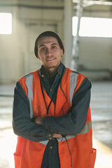 Leinwandbilder - Happy young engineer or foreman in protective fluorescent vest looking at camera with his arms crossed by chest while standing in workshop