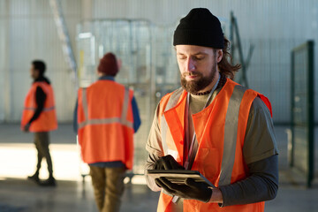 Leinwandbilder - Young bearded engineer in workwear scrolling in tablet in front of camera while standing in warehouse against two colleagues or subordinates