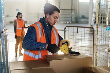 Poster - Side view of young male worker of warehouse in reflective vest sealing large cardboard boxes on cart with cellotape while bending over stack