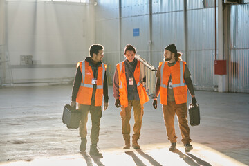 Poster - Three young intercultural engineers in workwear moving along warehouse while two of them with toolboxes looking at colleague with stepladder