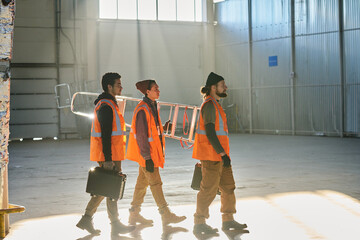 Leinwandbilder - Side view of three young engineers or technicians of warehouse carrying stepladder and toolboxes while moving along spacious workshop
