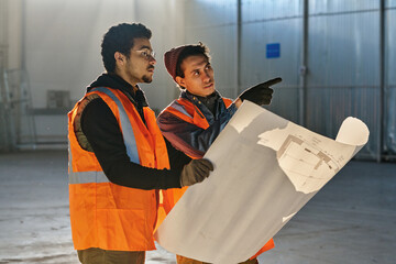 One of two engineers pointing forwards while explaining points of sketch to his colleague holding big blueprint at working meeting in warehouse