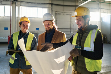 Leinwandbilder - Young smiling engineer pointing at building plan in blueprint while explaining it to contractor and colleague at meeting in warehouse