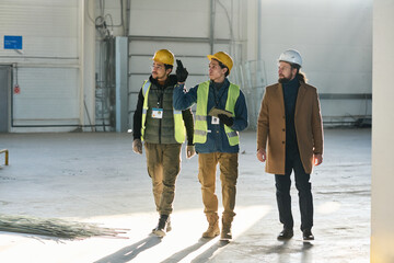 Leinwandbilder - Young confident engineer with tablet pointing at industrial equipment or machine while moving along spacious warehouse between colleagues