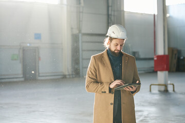 Leinwandbilder - Young bearded builder or solopreneur in hardhat scrolling in tablet while standing in front of camera in spacious sunlit warehouse and networking