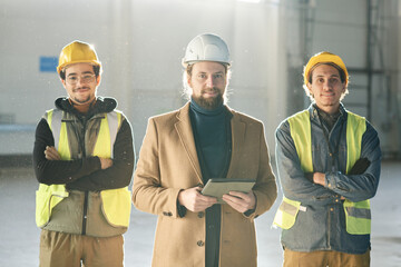 Leinwandbilder - Young successful foreman with tablet standing between two engineers in hardhats and protective fluorescent vests and looking at camera