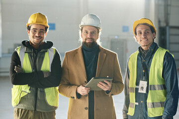 Leinwandbilder - Three happy young intercultural engineers in protective helmets looking at camera with smiles while two men in safety vests surrounding foreman