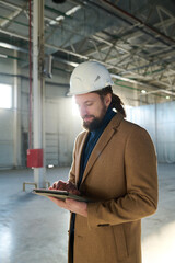 Leinwandbilder - Young serious contractor looking through online information on tablet screen while standing in front of camera at construction site