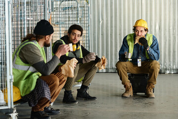 Leinwandbilder - Two young multiethnic engineers looking at their colleague duaring discussion of working points and eating sandwiches with hot tea