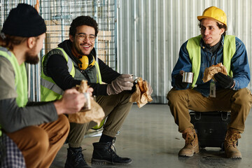 Happy young engineer with sandwich and cup of hot tea chatting to colleagues at lunch break while sitting in front of them in warehouse