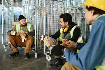 Leinwandbilder - Focus on two young intercultural engineers with sandwiches and cups of tea discussing latest news at lunch break while sitting on large carts
