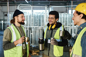 Leinwandbilder - Three happy young workers with metallic mugs of hot tea having chat while standing in front of camera against large carts in warehouse
