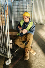 Poster - Young multiethnic male engineer in protetive workwear scrolling in mobile phone while sitting on large metallic cart and having tea at break