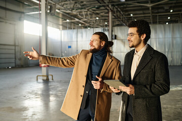 Leinwandbilder - Young confident businessman in coat stretching arm forwards while showing his colleague area for new office in unfinished building