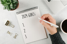 Woman's Hand Writing 2024 Goals In Notebook. Laptop, Home Plant, Cup Of Coffee On Light Table. Top View, Flat Lay