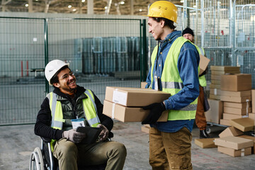 Leinwandbilder - Happy young engineer in wheelchair looking at colleague carrying stack of packed cardboard boxes while talking to him during work in warehouse