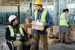 © pressmaster - Young engineer with stack of packed cardboard boxes talking to colleague sitting in wheelchair and using tablet during discussion of working points