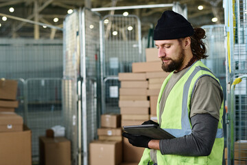 Leinwandbilder - Side view of bearded worker of warehouse in reflective vest looking through database in tablet while making revision of packed goods