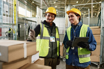 Leinwandbilder - Happy young man in workwear using tablet and listening to colleague pointing at stack of packed boxes on metallic cart during work
