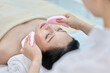 아시아 한국의 젊은 여성이 스파 또는 피부관리전문점 또는 피부과 병원의 침대에 누워 베이지색 대형 타올을 덮고 눈을 감고있고 하얀색 셔츠를 입은 전문관리사가 손에 분홍색 클렌징 스펀지를 들고 얼굴 피부관리를 하고 있다. 