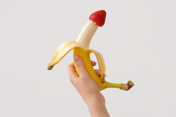 Female hand with fresh banana and strawberry on light background, closeup. Sex education concept