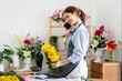 © Katcha - Female florist in apron using smartphone to talking receive order with customer while holding yellow