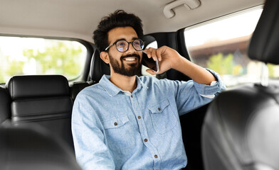 Wall Mural - Smiling young indian man talking over phone, sitting in taxi