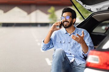 Wall Mural - Excited guy sitting in open car trunk, have phone conversation