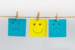 Positive attitude concept, three sticky notes hanging on a string attached with wooden clothespins, two sad faces in blue color and one standing out smiling face in yellow color in the middle.

