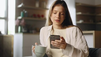 Charming young woman with cup of hot coffee or tea hold smartphone browsing scrolling app watching social media feed indoors Happy female texting on her phone enjoying leisure time at cozy home