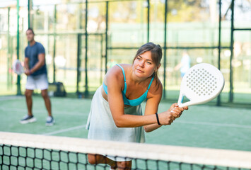 Wall Mural - Portrait of sporty young woman hitting two handed backhand during paddle tennis friendly match in open court on summer day..