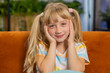 Close-up portrait of happy smiling Caucasian preteen school girl 7 years old. Young blonde lovely child kid looking at camera at home play room apartment sitting on orange couch. Ponytails hairstyle