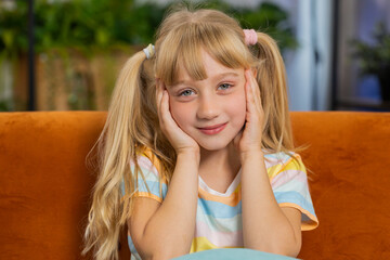 Close-up portrait of happy smiling Caucasian preteen school girl 7 years old. Young blonde lovely child kid looking at camera at home play room apartment sitting on orange couch. Ponytails hairstyle